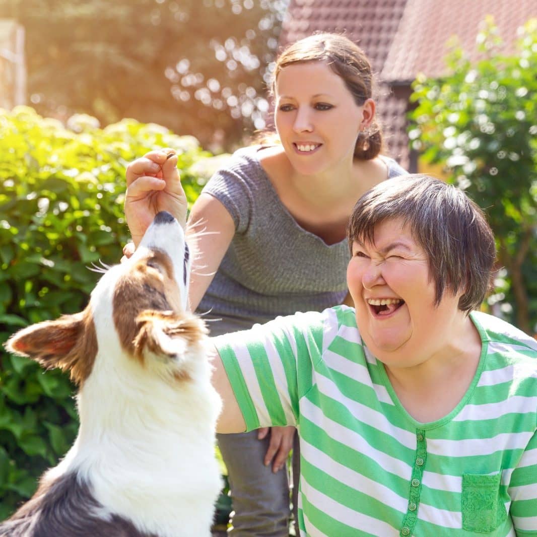 Two women in the garden playing with a white and brown dog. They are both laughing and having a good time.