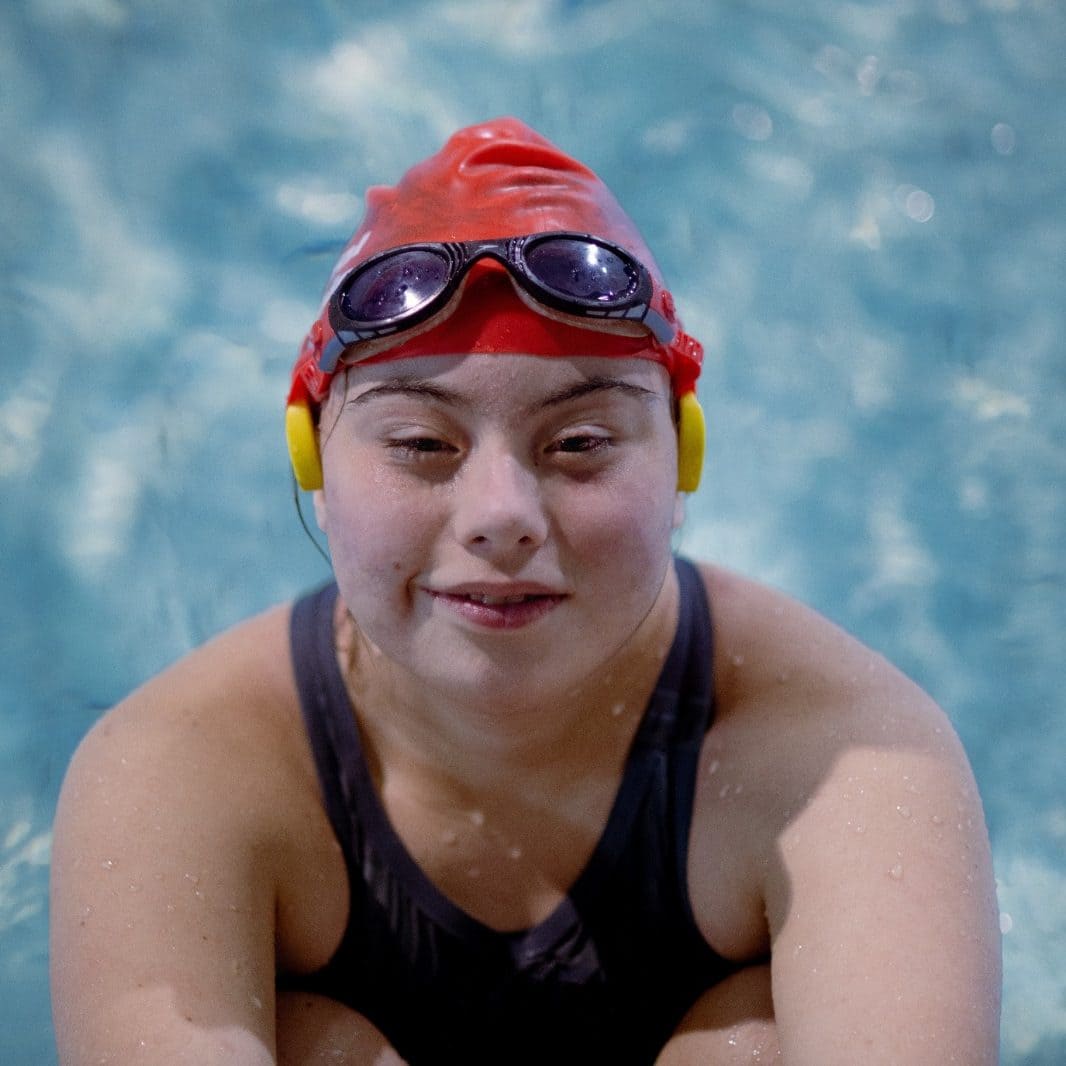 A woman in a swimming pool taking a break from swimming. She is looking up at the camera and smiling.