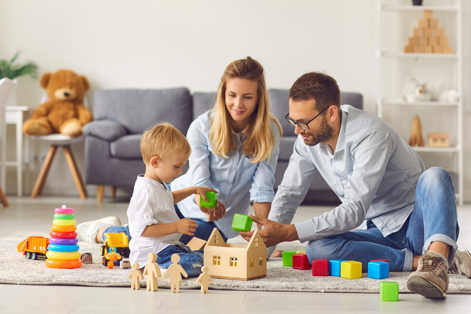 Parents playing with their young son on a rug. There are coloured blocks, trucks, and wooden toys.