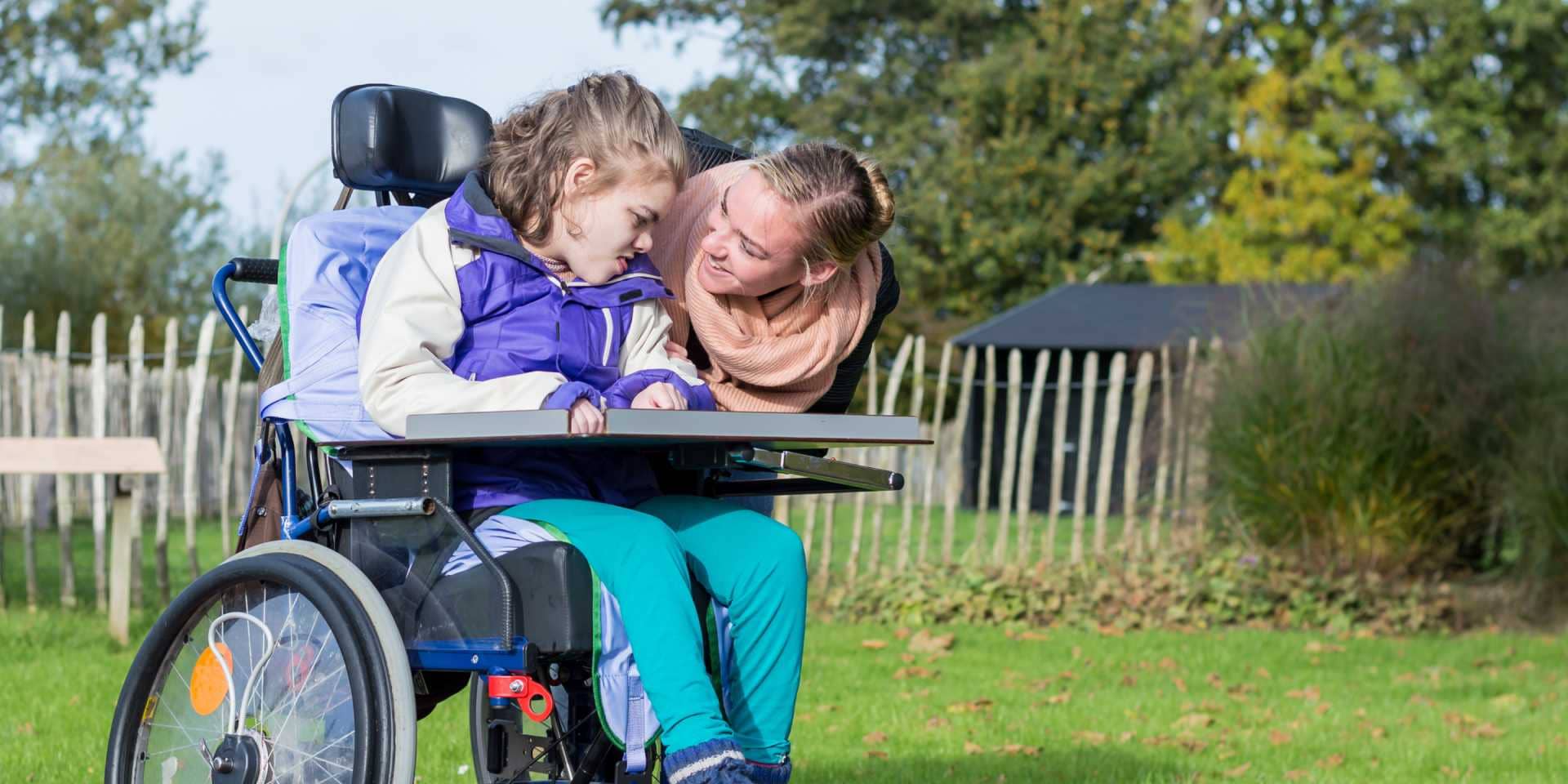 A support worker leans in to talk to a young girl who is a wheelchair user.