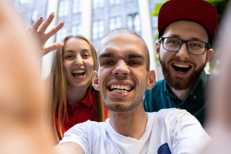 A man takes a group selfie on the street with his two friends on either side. They are all smiling and laughing as they pose for the photo.