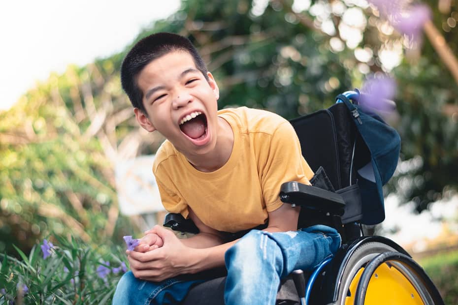 A boy with black hair is sitting in the garden. He is leaning forward in his wheelchair and laughing.