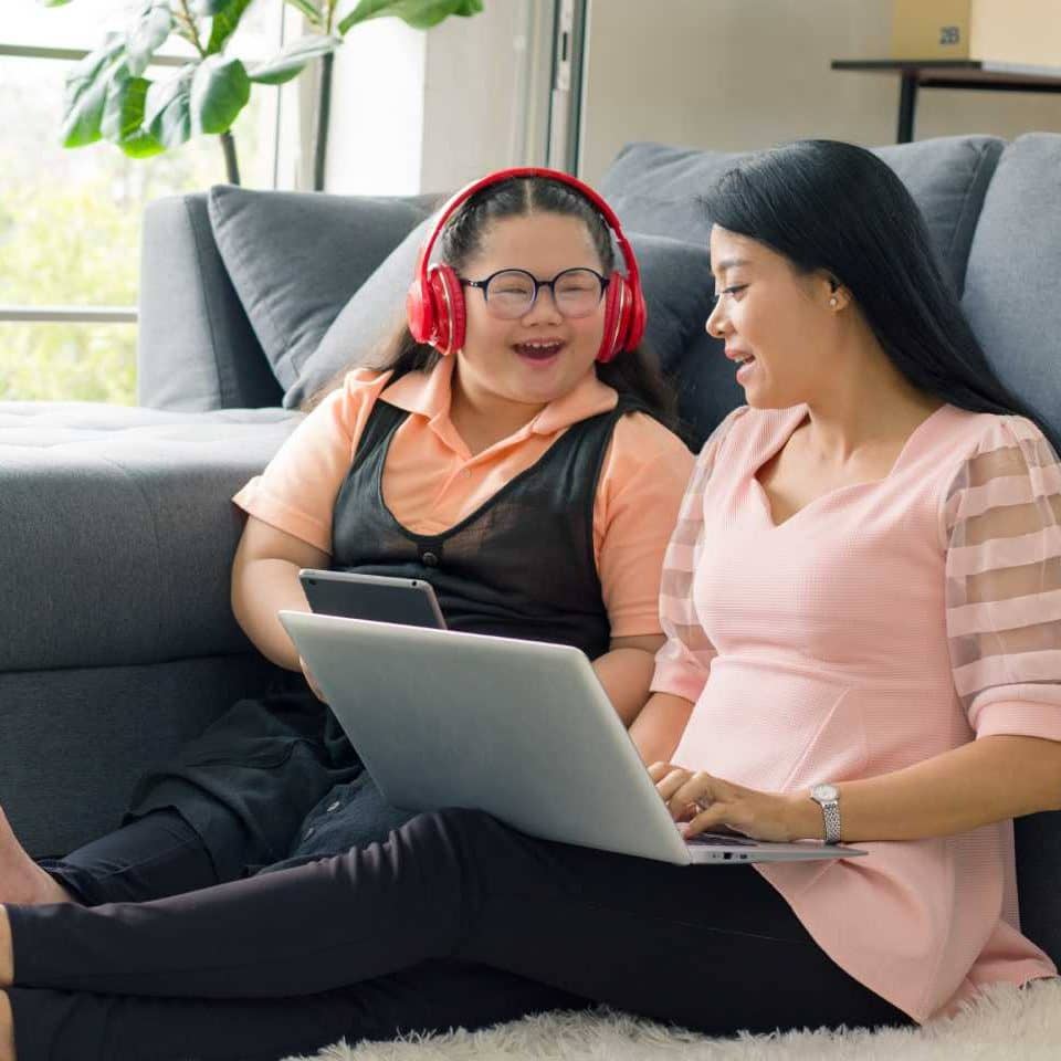 A young woman wearing headphones and glasses is smiling and engaging with another woman. Both are using devices and are sitting on the floor against a couch.